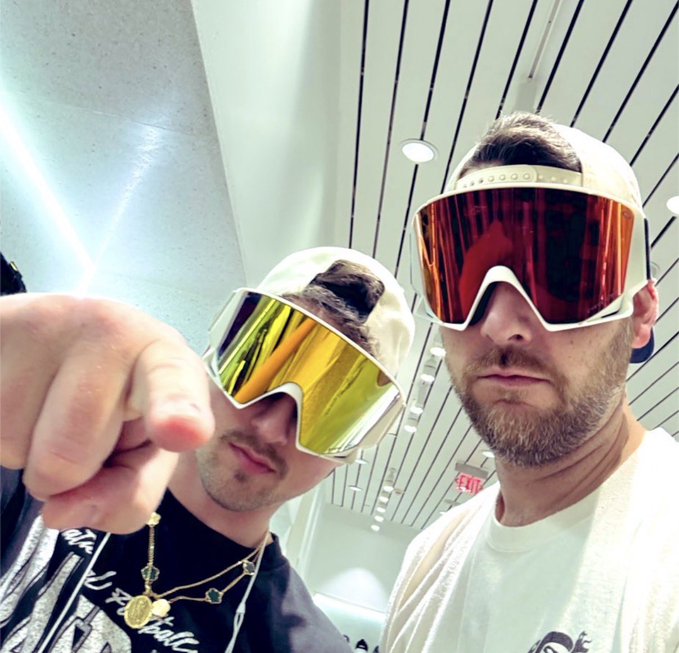 A selfie of two men wearing large ski goggles with reflective lenses, one yellow and one red/orange.