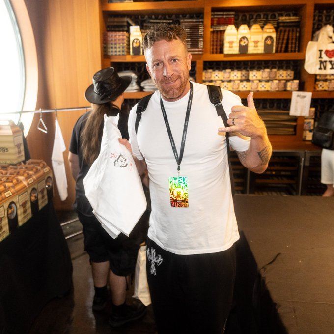 A man wearing a white t-shirt and a colorful NFT event lanyard holds up merchandise bags and makes a hand gesture while standing in what appears to be a shop or event venue.