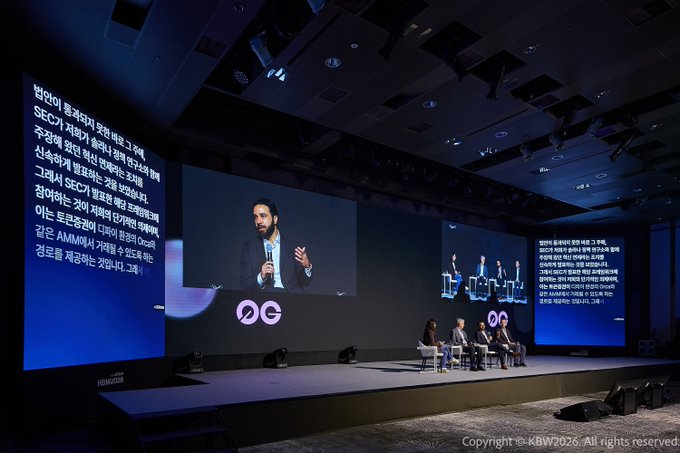 A conference stage (KBW 2026) featuring a speaker displayed on large screens with Korean text about the SEC and regulation of stablecoins including mentions of USD1, Ondo, Tether, Circle, and AMINA, with a panel seated on stage.