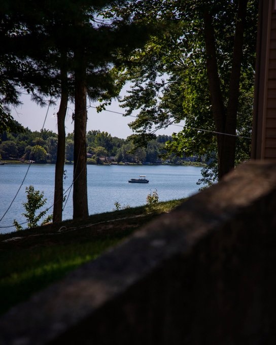 A serene lakeside view framed by tall pine trees, with a small boat floating on the calm blue water and a wooden railing in the foreground.