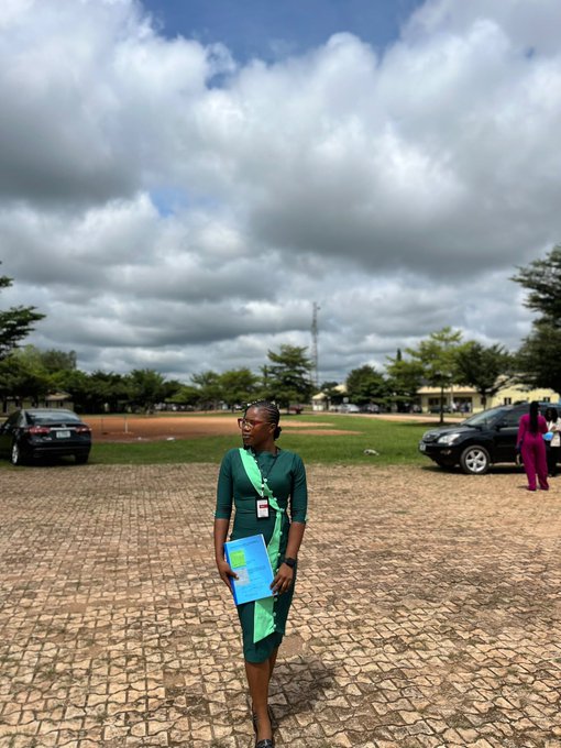 A woman wearing a green dress with a sash stands outdoors on a brick-paved area holding a blue folder, with a cloudy sky, parked cars, and trees in the background.