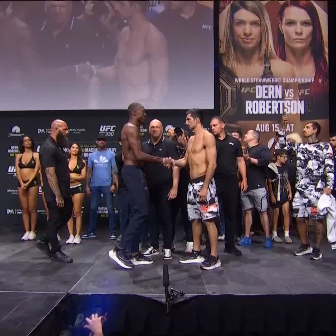 Two fighters, Jalin Turner and Kaue Fernandes, shake hands and stare each other down face-to-face during the UFC 330 weigh-in, with a "World Strawweight Championship: Dern vs Robertson, Aug 15" promotional poster visible on the large screen behind them.