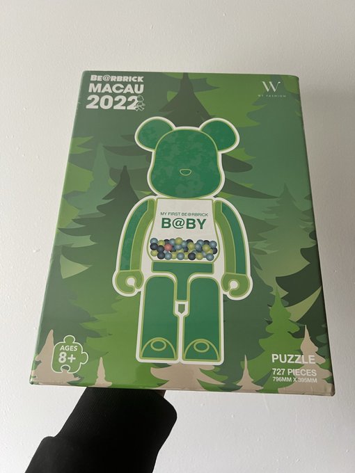 A hand holds a green Bearbrick x Macau 2022 puzzle box (727 pieces, 796mm x 396mm) featuring a transparent green bear figure against a forest background, marked 'MY FIRST BE@RBRICK B@BY' and 'AGES 8+'.