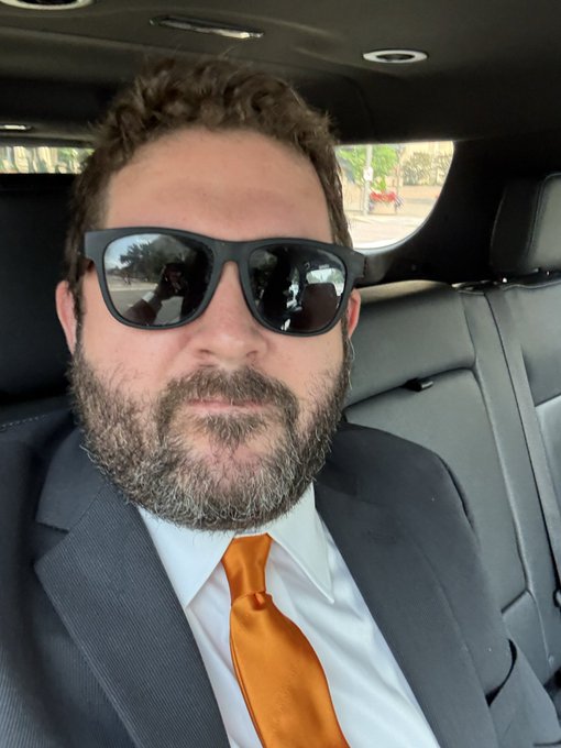 A bearded man wearing sunglasses, a gray suit, white shirt, and bright orange tie sits in the back seat of a car, projecting a confident "Hero" vibe as he prepares to take on the day.