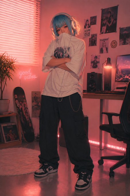 A cosplayer with short blue hair and headphones standing in a pink-lit room with a 'good vibes' neon sign, posters, and a desk setup.