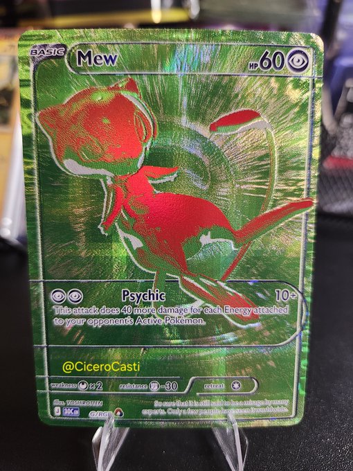 A shiny green holographic Mew Pokémon trading card from the Surging Sparks set, marked 'BASIC' with 60 HP and the 'Psychic' attack, displayed on a stand with the handle '@CiceroCast1' visible.