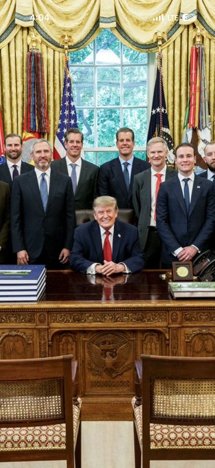 President Donald Trump sits smiling at the Resolute Desk in the Oval Office, surrounded by a group of executives standing behind him, with "2:00" visible on the clock atop the curtained windows. The image illustrates the post's triumphant theme—showcasing the CEO's hard-fought presence alongside the President as a symbol of victory after costly legal battles with the SEC and recognition as a leader in blockchain and payments.