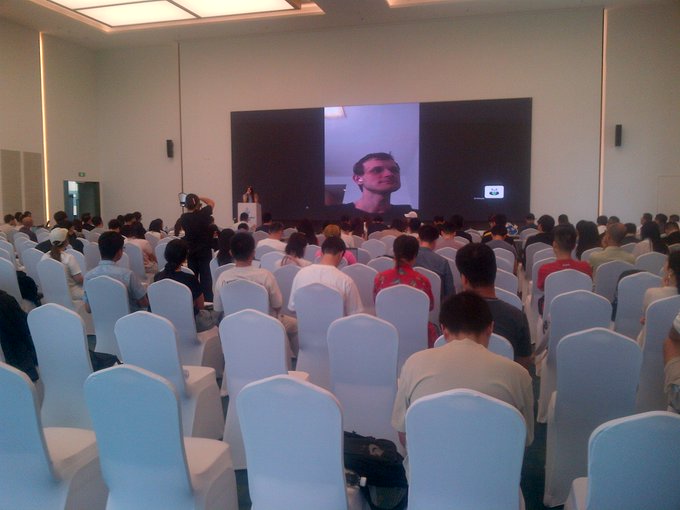 An audience sits in white-covered chairs watching a presentation, with a large screen displaying a man's face at the front of the room.