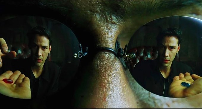 A man peers through magnifying lenses, with his reflection revealing a red pill in one hand and a blue pill in the other.