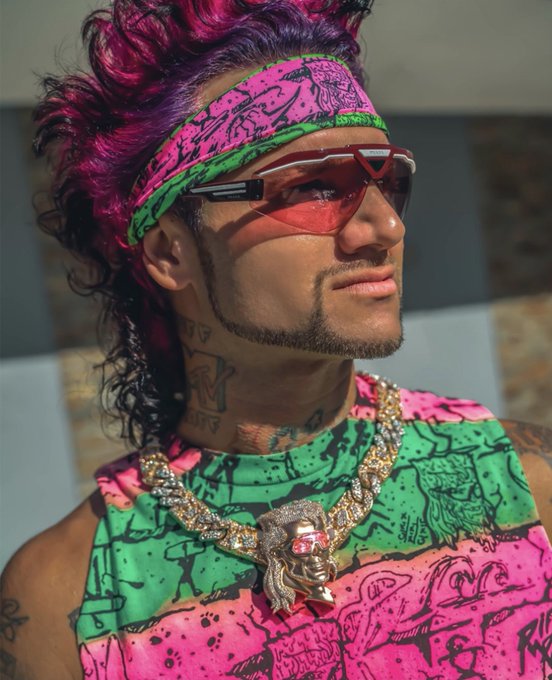 A man with vibrant pink and purple hair wears red-tinted wraparound sunglasses, a pink and green patterned headband, a matching tank top, and a large gold pendant necklace featuring a face with red eyes.