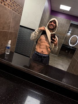 A mirror selfie taken in a public restroom shows a fit man with a beard wearing a gray hoodie pulled back to reveal his abs and gray sweatpants, holding a phone to take the photo.