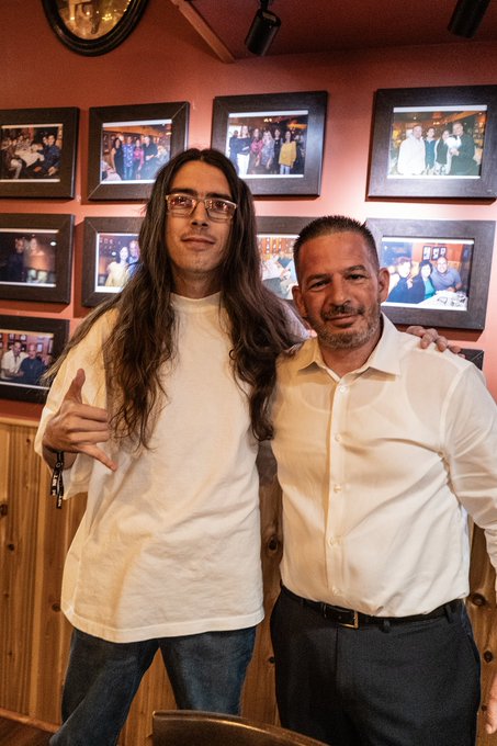 Two men pose together at an Alpha dinner, with a smiling bald man in a white shirt and belt standing beside a long-haired man with glasses in a white t-shirt making a hand gesture, both smiling warmly in front of a wall displaying framed photos of groups and event moments.