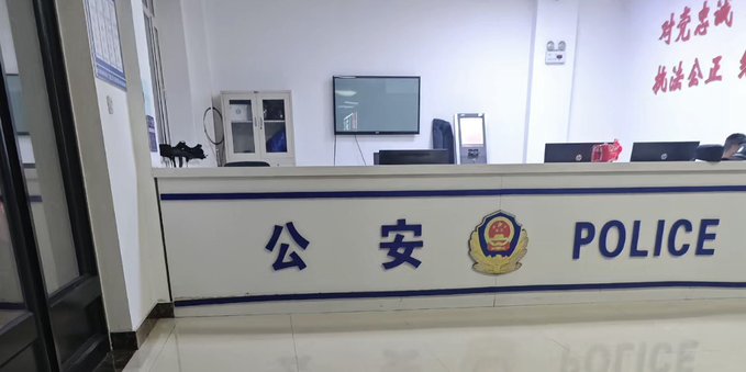 An interior view of a police station reception area features a white desk with blue text reading "公安 POLICE" and the official emblem, set against a white wall with red Chinese characters.
