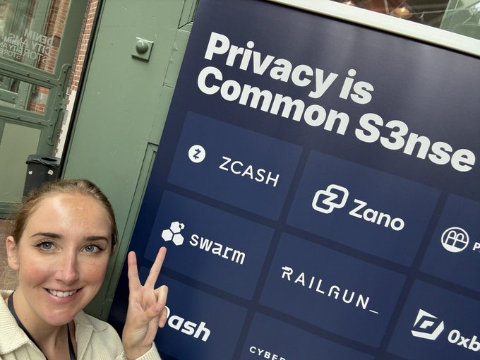 A woman smiling and making a peace sign while posing next to a display that reads 'Privacy is Common Sense' featuring logos for ZCASH, Zano, Swarm, Railgun, and others.