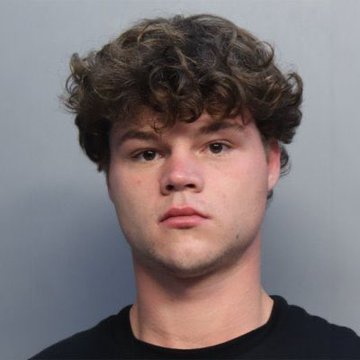 A mugshot-style portrait of a young man with curly brown hair wearing a black t-shirt, photographed against a plain gray wall.
