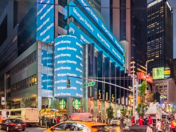 A Morgan Stanley building in a bustling city is wrapped in illuminated blue signage displaying the firm's name, visually anchoring the headline about the bank's executives predicting an end to traditional 9-to-5 banking amid the rise of tokenization and 24/7 trading.