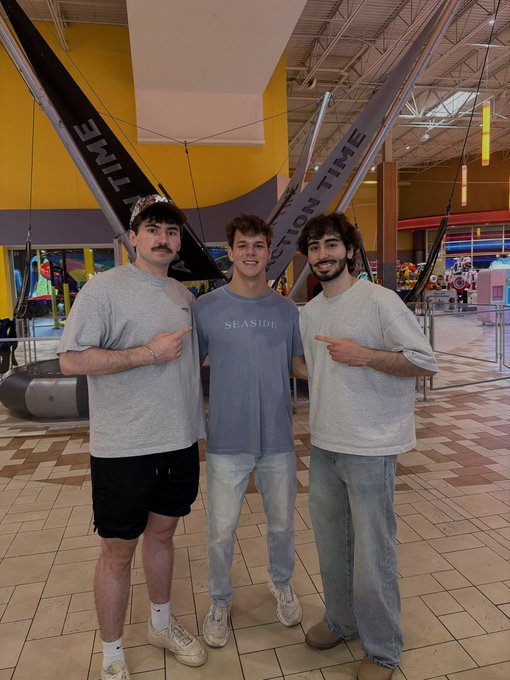 Three young men pose together inside what appears to be a trampoline or family entertainment center, with the middle person pointing to the friend on the right who wears a "SEASIDE" t-shirt. Behind them, large banners display partial text reading "STAY" and "ATION TIME," along with arcade games and a bungee attraction visible in the background, suggesting a casual, fun outing that aligns with the post's theme of "onboarding the masses" to the tagged individuals.
