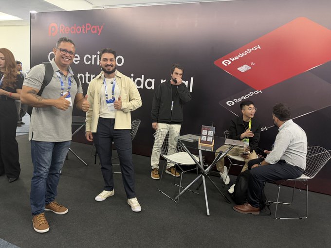Two men pose giving thumbs up at a 'RedotPay' booth while others work on laptops nearby at what appears to be a trade show.