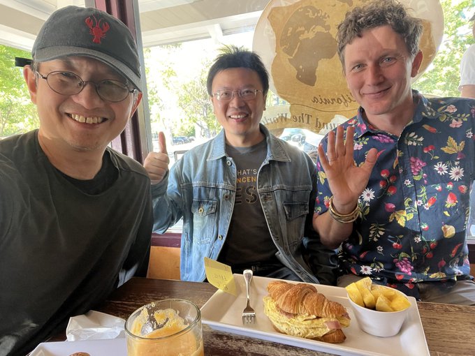 Three men are smiling and posing for a photo at a restaurant table that has a tray with a croissant sandwich, a side of potatoes, and a glass of orange juice.