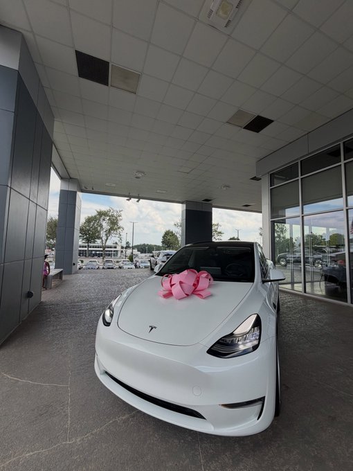 A brand new white Tesla car parked under a dealership awning, decorated with a large pink bow on the hood.