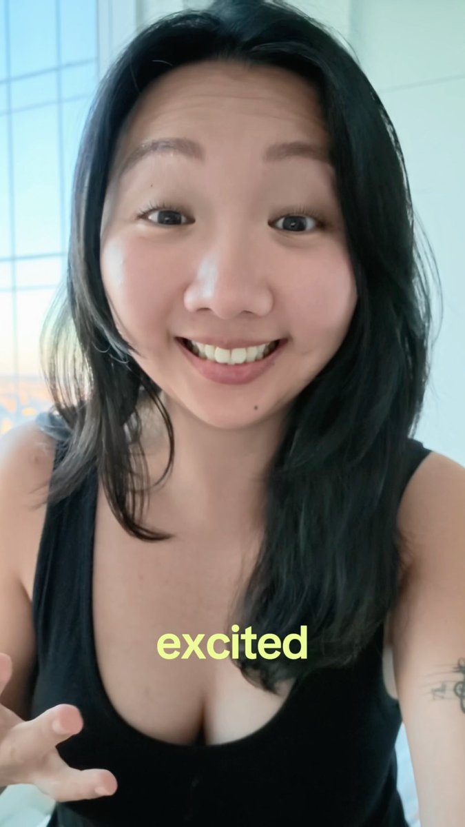 A woman with long black hair wearing a black top smiles at the camera in a selfie-style video, with the word 'excited' displayed in yellow text.