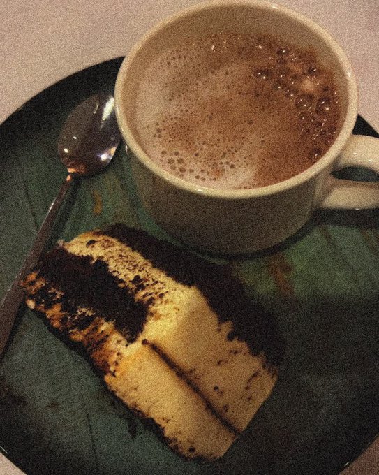 A mug of frothy hot chocolate sits on a green plate next to a slice of layered chocolate and vanilla cake, with a spoon resting beside it.