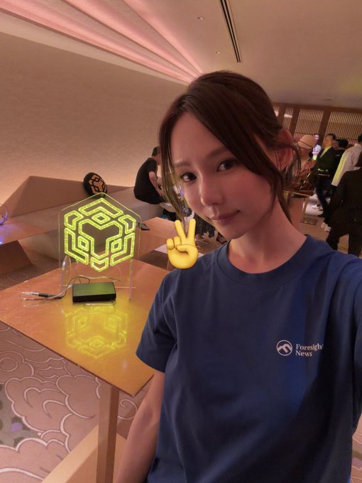 A young woman wearing a "Foresight News" t-shirt poses with a peace sign next to an illuminated BNB Chain hexagonal logo at a lively BNB Chain event. The scene captures the vibrant atmosphere she described, with attendees mingling in the background of the venue.