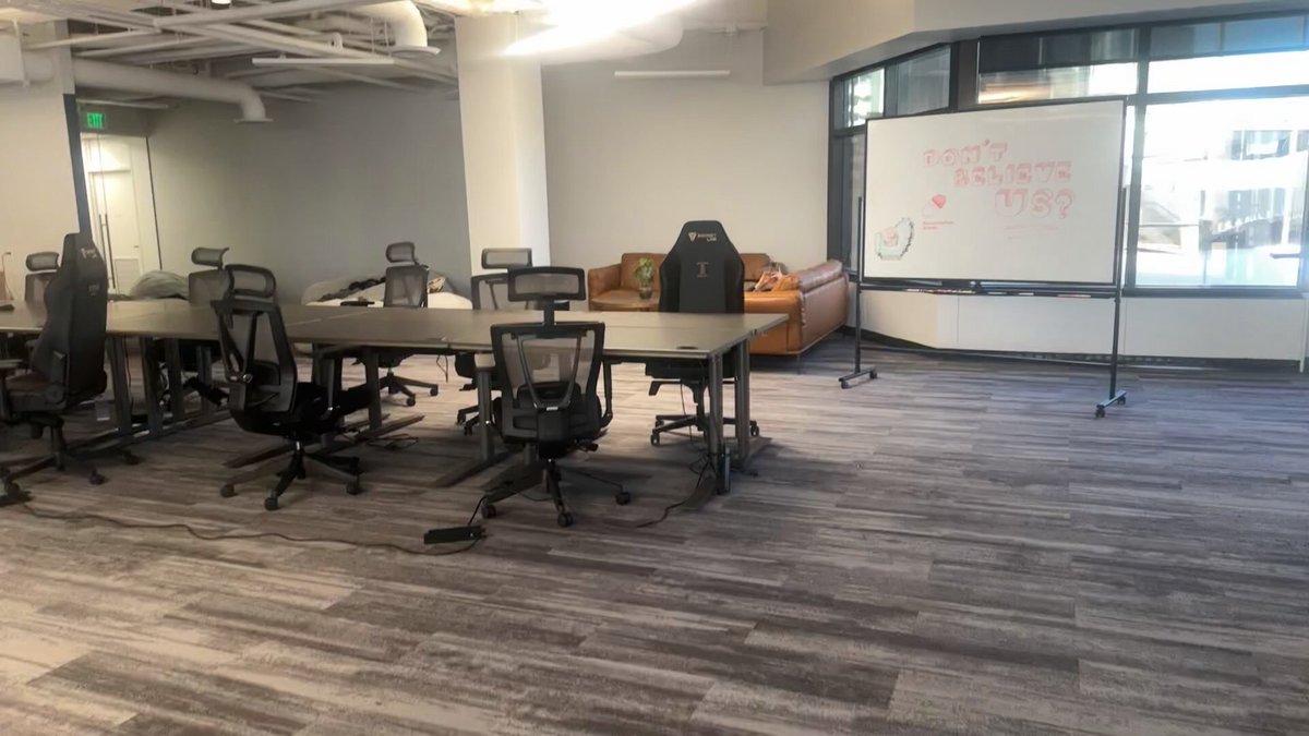The image shows an office space featuring gray carpeting, multiple tables and chairs, and a whiteboard with writing on it. The room appears to be designed for collaboration or meetings, as there are several people present in the scene. A couch is also visible within the room, providing additional seating options. The post about the cheat code seems unrelated to the office setting but could potentially be related to an event or gathering taking place at this location.