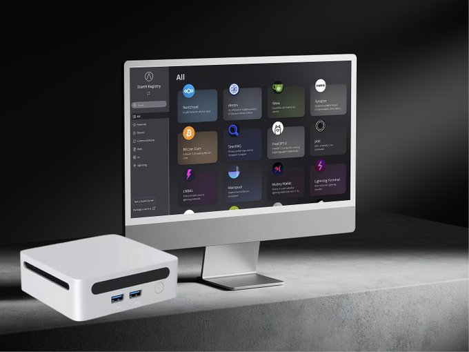 A small white desktop computer (Mini PC) sits next to a monitor displaying a 'Start Registry' interface with various app icons like NextDNS and denys.