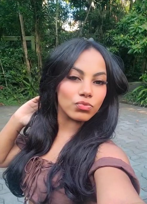 A selfie of a woman with long dark wavy hair, wearing a brown top, standing outdoors with green foliage in the background.