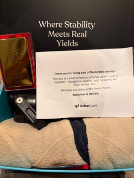 An open box containing a gold-colored item and a thank you card reading 'Where Stability Meets Real Yields - Thank you for being part of the Unisaks journey. We hope you enjoy every piece inside. Welcome to Unisaks.'