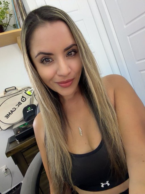 A woman with long blonde-highlighted hair wearing a black Under Armour top and a feather pendant necklace, taking a photo in front of a white door with a 'BCGWME' bag visible behind her.