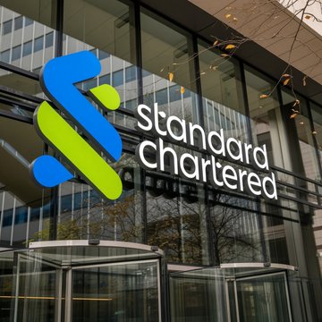 The Standard Chartered bank logo is displayed on the entrance of a glass-fronted building, featuring the company's distinctive blue and green interwoven emblem alongside its name. The image contrasts with the accompanying post's discussion of blockchain investments like $LINK, as Standard Chartered represents traditional banking rather than cryptocurrency, highlighting the gap between conventional finance and emerging digital asset investment opportunities.
