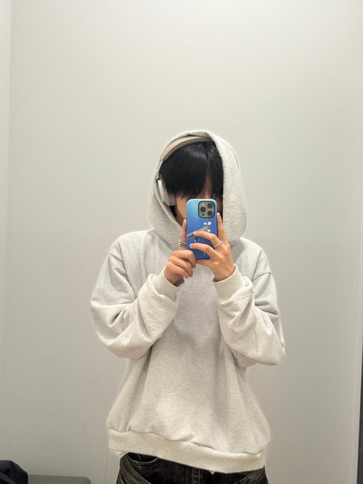 A mirror selfie of a person wearing a gray hoodie pulled up over their face and white headphones, holding a blue smartphone.