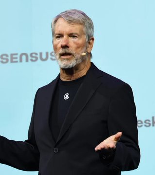 An older man with grey hair and beard wearing a black blazer and shirt, speaking on stage with a microphone headset, with 'SENSUS' visible in the background.