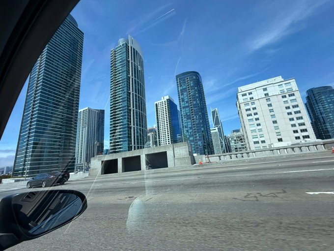 A view from inside a car shows a sunny cityscape with tall skyscrapers, including glass-fronted residential towers and a beige hotel building under a blue sky with wispy clouds.