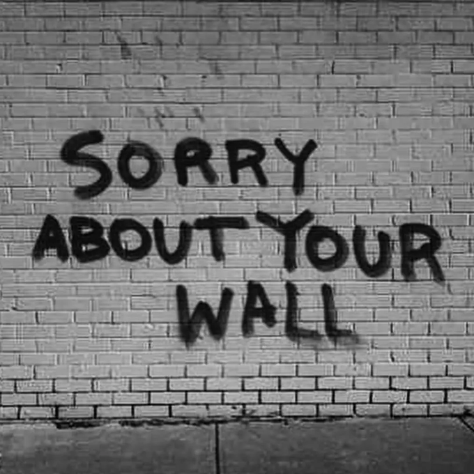 Black spray-painted graffiti on a white brick wall reads "SORRY ABOUT YOUR WALL."