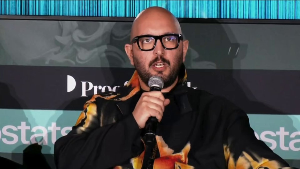 The image features a bald man wearing glasses, who appears to be speaking into a microphone at a podium. The background of the image shows a banner with the text "Products Estates" written on it. The main subject of the image is the bald man holding a microphone and talking about why the subnet could not exist without Bittensor.
