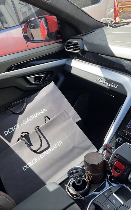 The interior of a Lamborghini (visible logo on the dashboard) with Dolce & Gabbana shopping bags and a Louis Vuitton cup placed in the passenger seat area.