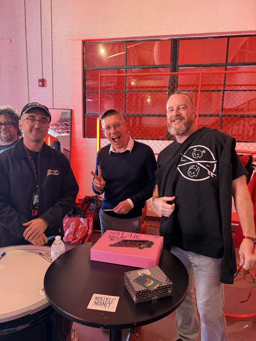 Three men posing happily behind a round black table holding a pink cake with '40' written on it and a 'UNION SALON' card, in a room with red-lit windows.