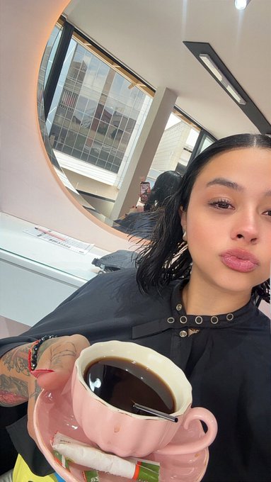 A woman with dark hair takes a selfie while holding a pink teacup filled with coffee, set in a salon with a round mirror behind her.