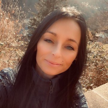 An outdoor selfie of a woman with long dark hair smiling, taken with a scenic background of trees and a valley.