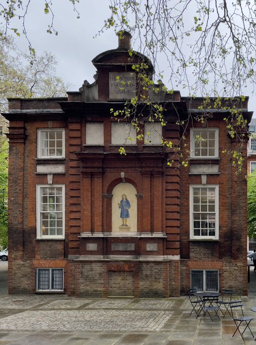 A historic red brick schoolhouse featuring a decorative niche with a figure in blue and an inscription reading "THE BLUE COAT SCHOOL FOUNDED 1689" stands behind budding spring branches, with café seating visible on the cobblestone courtyard in front.