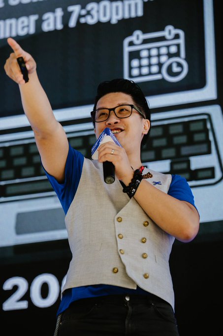 A man wearing glasses, a blue shirt, and a gray vest with gold buttons is speaking into a microphone and pointing, with '7:30pm' and a calendar icon visible on the screen behind him.