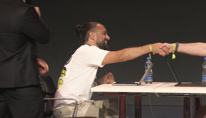 A bearded trader in a white t-shirt shakes hands with another competitor across a table lined with laptops, celebrating his victory at a trading competition with sponsors visible from Solflare, Oligarchxyz, and EyeKon Studio.