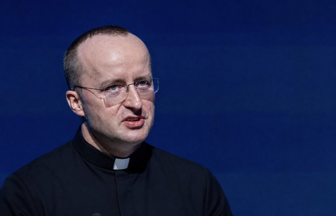 A clergyman wearing a black clerical shirt and glasses speaks at a podium against a blue background, delivering what appears to be a prayer-like message asking for "Our Father...Thy kingdom come" and for the cryptocurrency $CRO to "finally rise," humorously blending traditional religious devotion with crypto investment hopes.