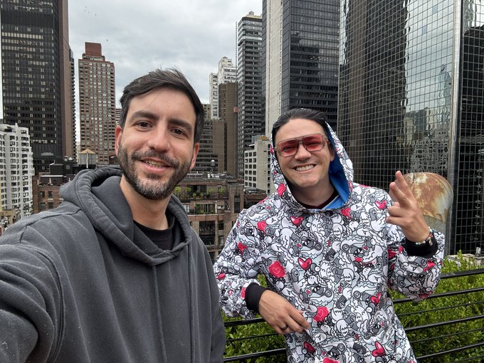 Two men taking a selfie in a city setting; one wears a gray hoodie while the other wears a colorful patterned jacket with red sunglasses and makes a hand gesture.