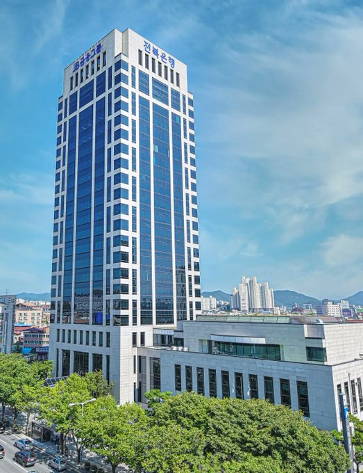 A tall modern office tower with Korean signage reading "전북은행" (Jeonbuk Bank) stands prominently against a blue sky, representing the headquarters of the South Korean commercial bank that has become the first in the country to lend to companies using Bitcoin as collateral.