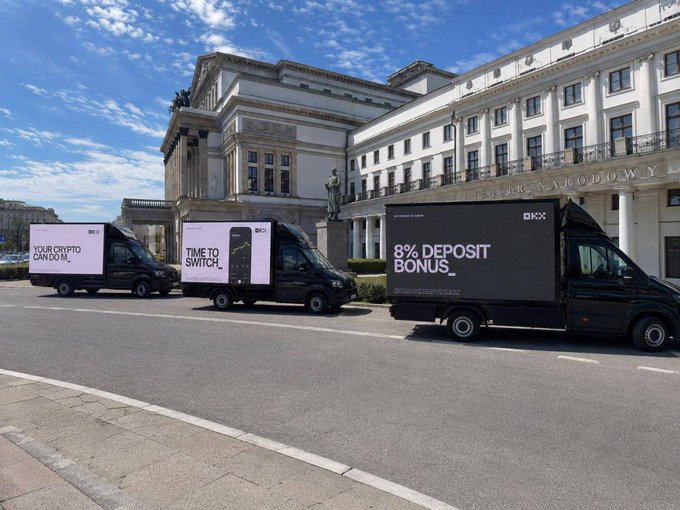 Three OKX-branded vehicles displaying digital billboard ads—including "YOUR CRYPTO CAN DO_M," "TIME TO SWITCH," and "8% DEPOSIT BONUS" at okx.com/okxpl—parked outside a classical European building, visually amplifying OKX's promotional push to attract new EU users with an 8% deposit bonus as MiCA regulations take effect.