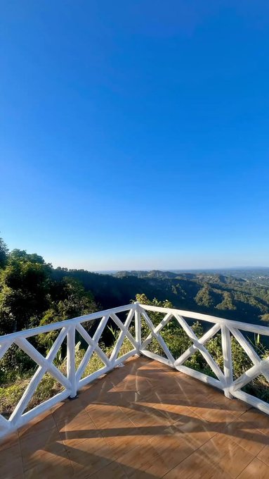 A scenic photograph from a white-railed balcony or terrace with tiled flooring, overlooking a vast green mountainous landscape under a clear blue sky.